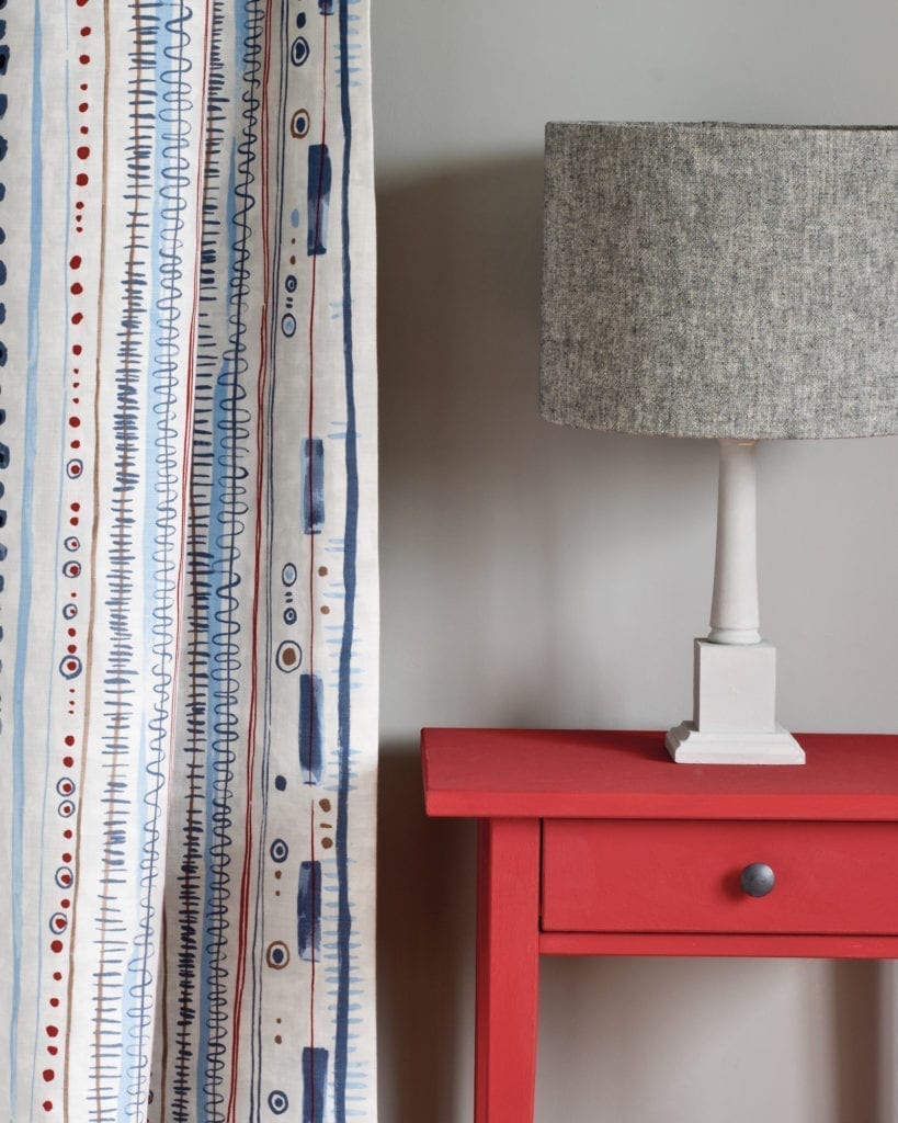 Side table painted with Chalk Paint® in Emperor's Silk, a bright pure red against a wall painted with Paris Grey. Curtain in Piano in Old Violet and lampshade in Linen Union in Graphite + Old White