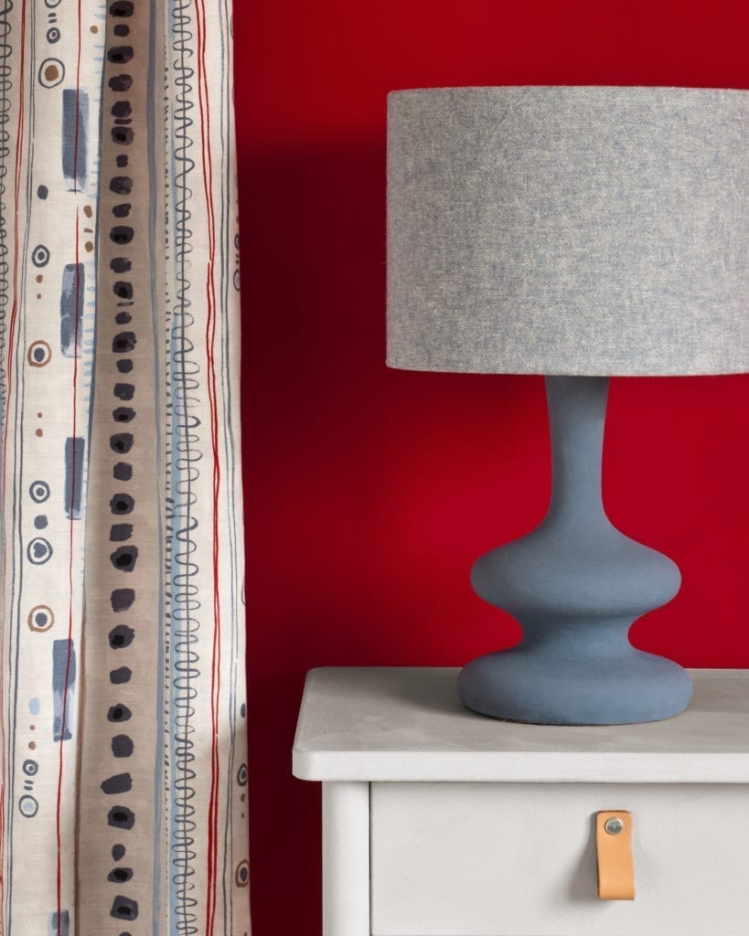 Side table painted with Chalk Paint® in Chicago Grey, a cool, fresh and modern grey, with a hint of blue against a red wall painted with Emperor's Silk. Curtain in Piano in Old Violet and lampshade in Linen Union in Old Violet + Old White