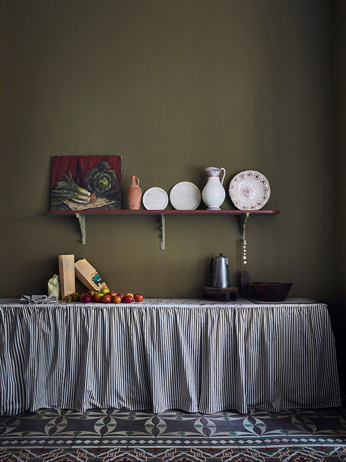 A kitchen painted in Annie Sloan Olive Wall Paint, with a dining table covered in striped fabric and dishes and mugs placed on a shelf along the wall.