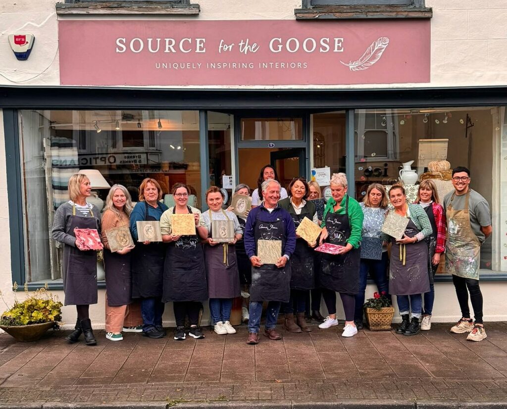 a group of people holding painted boards outside a shop called Source for the Goose